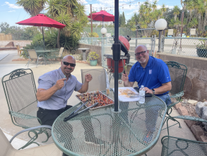 two men sitting at a patio table outdoors setting up some bbq snacks