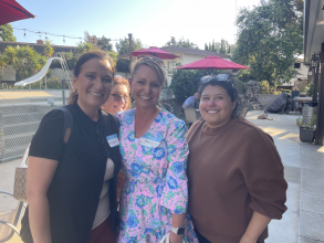 Three women standing and smiling together in an outdoor patio with red umbrellas behind them