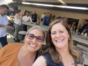 Two women standing together on an outdoor patio one woman has light hair and a rust colored shirt the other woman has brown hair and is in a dark outfit