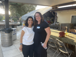 Two women standing together on an outdoor patio one woman has dark hair and a white shirt the other woman has brown hair and is in a black outfit